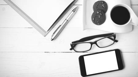 coffee cup with cookie,phone,notebook and eyeglasses on white wood table black and white colorの写真素材