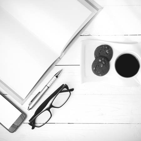 coffee cup with cookie,phone,notebook and eyeglasses on white wood table black and white color