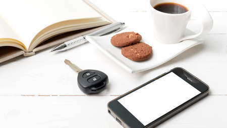 coffee cup with cookie,phone,open notebook and car key on white wood table