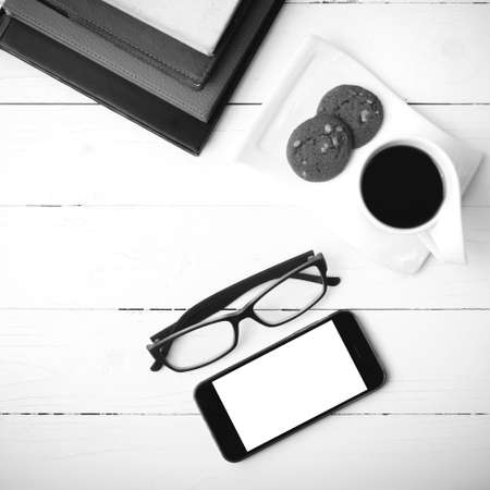 coffee cup with cookie,phone,stack of book and eyeglasses on white wood table black and white colorの写真素材