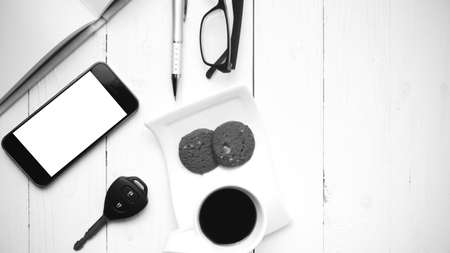 coffee cup with cookie,phone,open notebook,car key and eyeglasses on white wood table black and white colorの写真素材