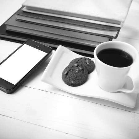 coffee cup with cookie,phone,stack of book and eyeglasses on white wood table black and white color