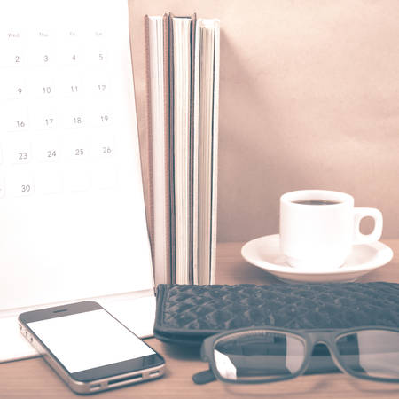 office desk : coffee with phone,stack of book,eyeglasses,wallet,calendar on wood background vintage styleの写真素材