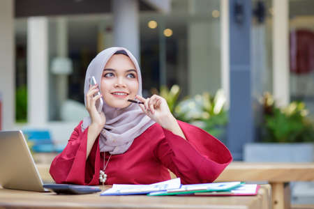 Young asian muslim woman working on laptop and talking on cell phone outdoor