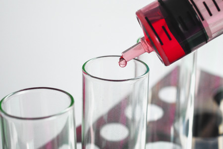 Syringe dropping a chemical solution into a test tube on soft light background.