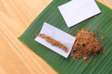 Homemade dry tobacco and cigarette over banana leave on the brown wooden table.