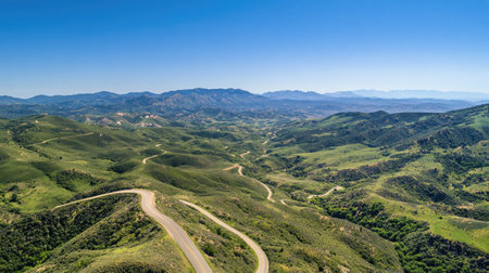 Aerial view of a winding road cutting through a mountainous landscape, green valleys below, and ample copy space in the clear blue sky.の素材