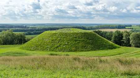 A serene view of an ancient burial mound amidst a lush green landscape under a partly cloudy sky. The site reflects rich history and natural beauty.の素材