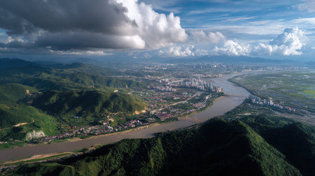 Captivating aerial perspective of a lush green landscape featuring a winding river, mountains, and an urban skyline under a dynamic sky filled with clouds.の素材