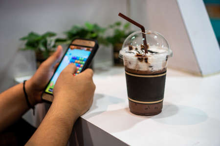 Cold Cocoa Drink And girls playing games on the mobile.Iced cocoa drink with whipping cream topping in cafeの写真素材