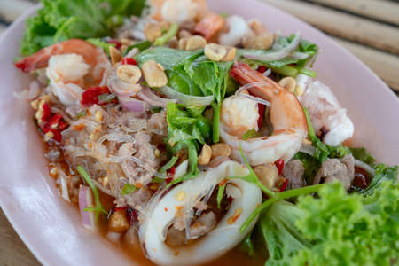 Spicy seafood salad is a Thai food for health consists of vermicelli, squid, shrimp, onion, chili and herb mixed in plate on a wood table at Thai street foodの写真素材