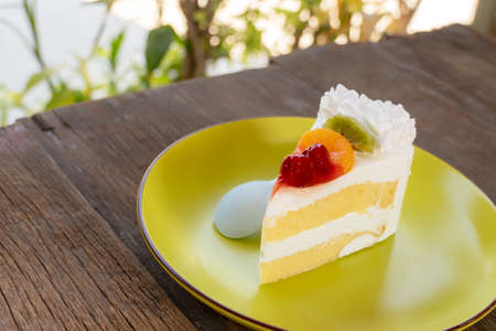 slice cream cake topped with fresh fruit such as grapes, oranges and kiwi in a green plate on a wooden tableの写真素材