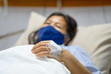 A sick Asian woman wear mask and lying on bed in hospital with IV saline drip to back of the hand. Corona Virus or Covid-19 concept.の写真素材