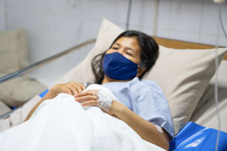 A sick Asian woman wear mask and lying on bed in hospital with IV saline drip to back of the hand. Corona Virus or Covid-19 concept.の写真素材