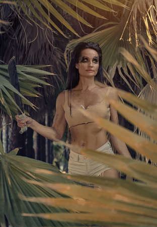 Middle aged young adult athletic sporty woman wear sexy short top and shorts holding machete posing alone on wild nature palm trees jungle forest. Spirit of adventure. Survivor amazon female conceptの写真素材