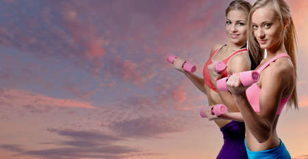 Two beautiful 20s women wear top and sporty pants doing fitness exercise with dumbbells looking at camera while pose on cloudy sky banner background with copy space. Wellness, sport lifestyle conceptの写真素材
