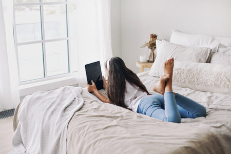 Adorable pre-teen 12s girl in wireless headphones lying on bed with digital tablet device. Young generation and modern tech overuse, gadget usage for fun, hobby and leisure on weekend at homeの写真素材