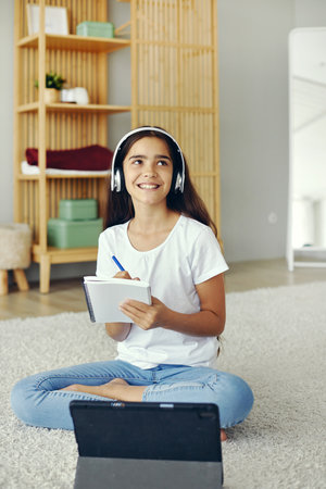 Pretty pre-teen girl sit on floor in cozy living room wear wireless headphones studying online use digital tablet device. Self-education, younger gen and modern tech usage for learning, gain new skillの写真素材
