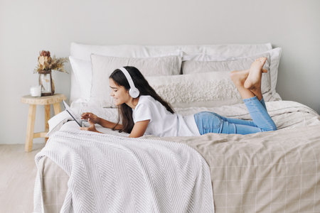 Adorable pre-teen girl in wireless headphones lying on bed with digital tablet device. Young generation and modern tech overuse, gadget usage for fun, hobby and leisure on weekend at homeの写真素材