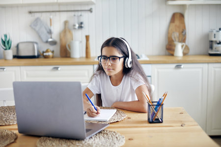 Pre-teen 12s girl in headphones and eyeglasses sit at table e-learns, listen online course, audio lesson, get new knowledge, skills using internet and modern tech. Development, education, eye-sightの写真素材