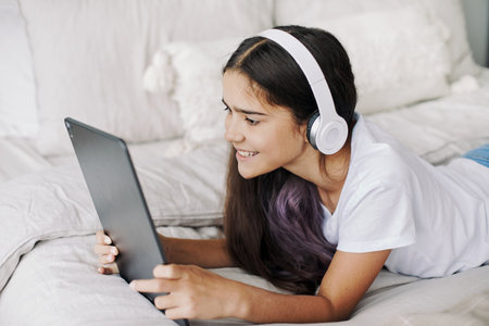 Adorable pre-teen girl in wireless headphones lying on bed with digital tablet device. Young generation and modern tech overuse, gadget usage for fun, hobby and leisure on weekend at homeの写真素材