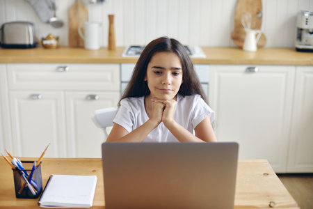 Pretty pre-teen 12s girl sit at table in kitchen watching on-line educational program, new videovlog or movie spend time on internet alone at home. Younger gen and modern tech usage for leisureの写真素材