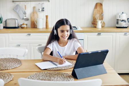 Pretty pre-teen 12s girl in wireless headphones sit at table e-learning, listen on-line course, audio lesson, receive new knowledge, skills using internet and modern tech. Child development. Educationの写真素材