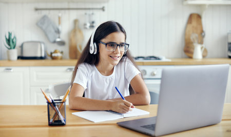 Pre-teen 12s girl in headphones and eyeglasses sit at table e-learns, listen online course, audio lesson, get new knowledge, skills using internet and modern tech. Development, education, eye-sightの写真素材