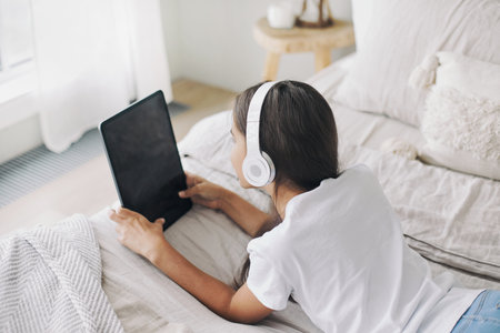 Adorable 12s pre-teen girl in wireless headphones lying on bed with digital tablet device. Young generation and modern tech overuse, gadget usage for fun, hobby and leisure on weekend at homeの写真素材