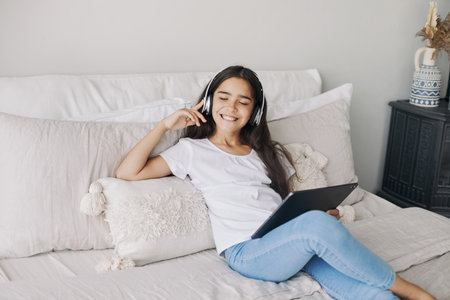 Cute pre-teen girl resting alone in bedroom with digital tablet device, wear wireless headphones enjoy favourite track, listen music on weekend at home. Young generation and tech usage, fun, leisureの写真素材