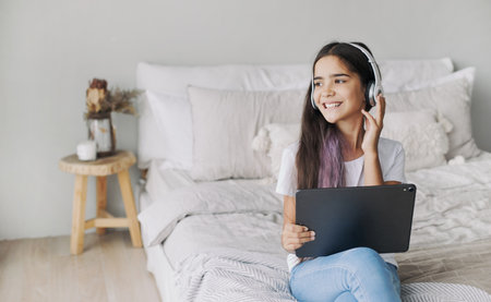 Cute pre-teen girl resting alone in bedroom with digital tablet device, wear wireless headphones enjoy favourite track, listen music on weekend at home. Young generation and tech usage, fun, leisureの写真素材