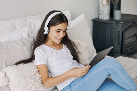Attractive pre-teen 12s girl wear wireless headphones using modern digital tablet seated on bed in cozy bedroom. I generation and technologies usage for fun, education and leisureの写真素材