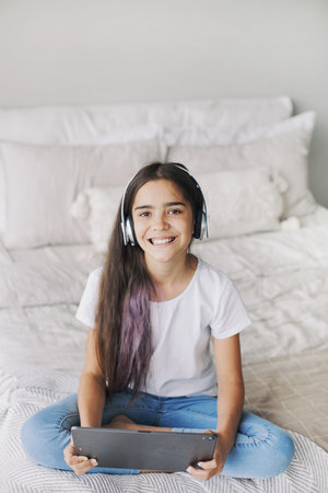 Attractive pre-teen 12s girl wear wireless headphones using modern digital tablet seated on bed in cozy bedroom. I generation and technologies usage for fun, education and leisureの写真素材