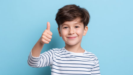 A joyful boy with a charming smile and short hair shows a thumbs-up gesture against a soothing blue background, radiating positivity and confidence.の素材