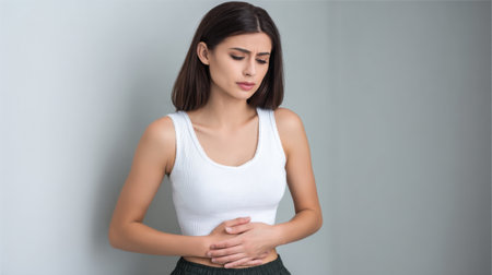 A young woman stands indoors, visibly distressed by stomach pain. She holds her abdomen, reflecting worry and discomfort in her expression, embodying everyday health challenges.の素材