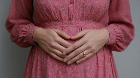 A serene moment captured of a pregnant woman in a pink dress, gently cradling her belly with hands forming a heart shape, symbolizing love and connection.の素材