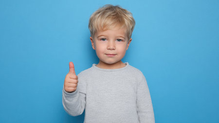 A young boy with blonde hair displays a thumbs up gesture against a bright blue background. His expression radiates joy and confidence, perfect for conveying positivity.の素材