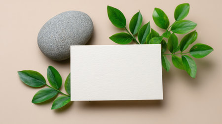 A serene and minimalist flat lay arrangement featuring a blank card, smooth stone, and fresh green leaves on a soft beige background, ideal for design use.の素材