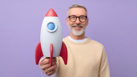 A cheerful senior man holds a colorful toy rocket, symbolizing dreams and aspirations, set against a vibrant purple background, radiating positivity.の素材