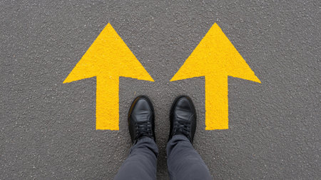 A top view of a person's feet wearing black shoes standing on gray pavement with two yellow arrows directing left and right, symbolizing decision-making.の素材