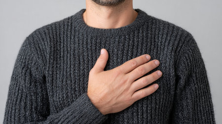 A man stands confidently with his hand placed over his heart, wearing a comfortable gray sweater. The image conveys warmth and sincerity in a neutral studio setting.の素材