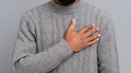 A man stands confidently with his hand placed over his heart, displaying a gesture of respect and sincerity. He wears a cozy gray sweater, embodying warmth and genuine emotion. The neutral background enhances the focus on his thoughtful expression.の素材