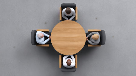 This image captures an aerial view of four individuals seated at a round wooden table, fostering collaboration and communication in a modern office setting.の素材
