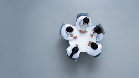 A top view of medical professionals in white coats engaged in a discussion around a round table, highlighting teamwork and collaboration in healthcare settings.の素材