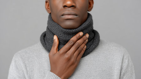 A thoughtful young man presents a serene expression while touching his neck, adorned in a gray sweater and matching scarf, in a minimalist setting.の素材