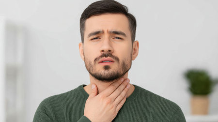 A young man displays discomfort as he touches his throat, reflecting emotions of worry or concern about his health in a modern indoor setting.の素材