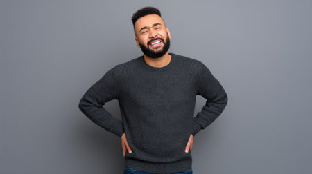 A young man stands against a gray background, exhibiting signs of back pain with a concerned expression. His casual attire suggests an everyday scenario, emphasizing the emotional and physical aspects of discomfort.の素材
