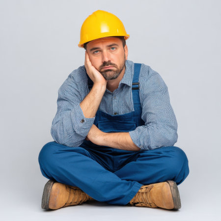 A construction worker sits on the floor against a gray backdrop, wearing a yellow hard hat and blue overalls. His thoughtful expression conveys a sense of contemplation and boredom, making this image ideal for themes of labor and industry.の素材