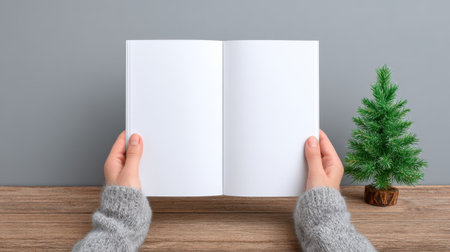A serene scene showcasing hands holding an open blank book against a cozy gray background with a small green Christmas tree beside a wooden table.の素材
