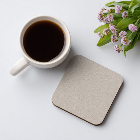 A serene and calming image featuring a white mug filled with steaming black coffee, a minimal square coaster, and a delicate flower bouquet, perfect for cozy mornings.の素材